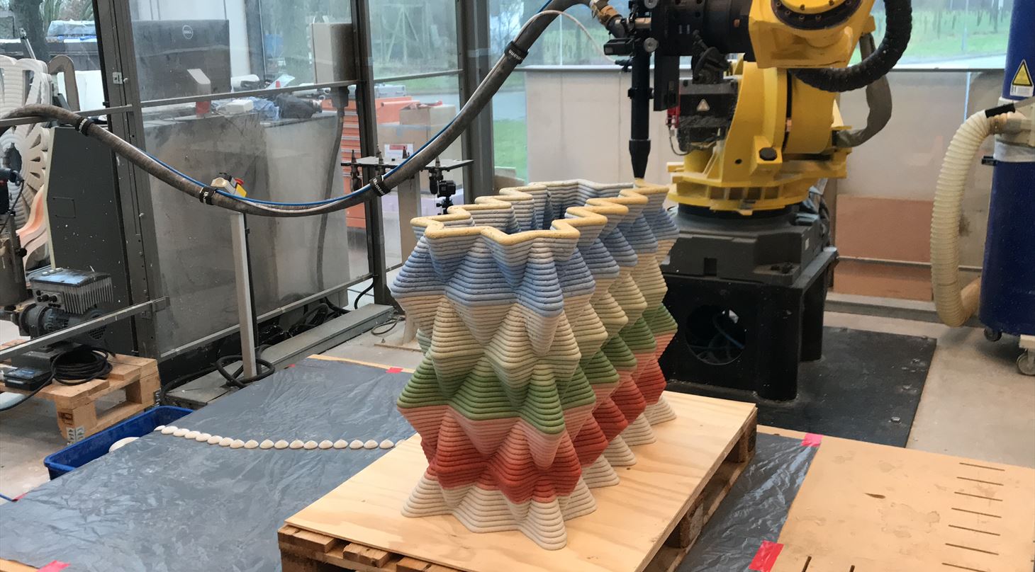 Billedet viser en robotarm, som er ved at 3D-printe en betonvæg i forskellige farver