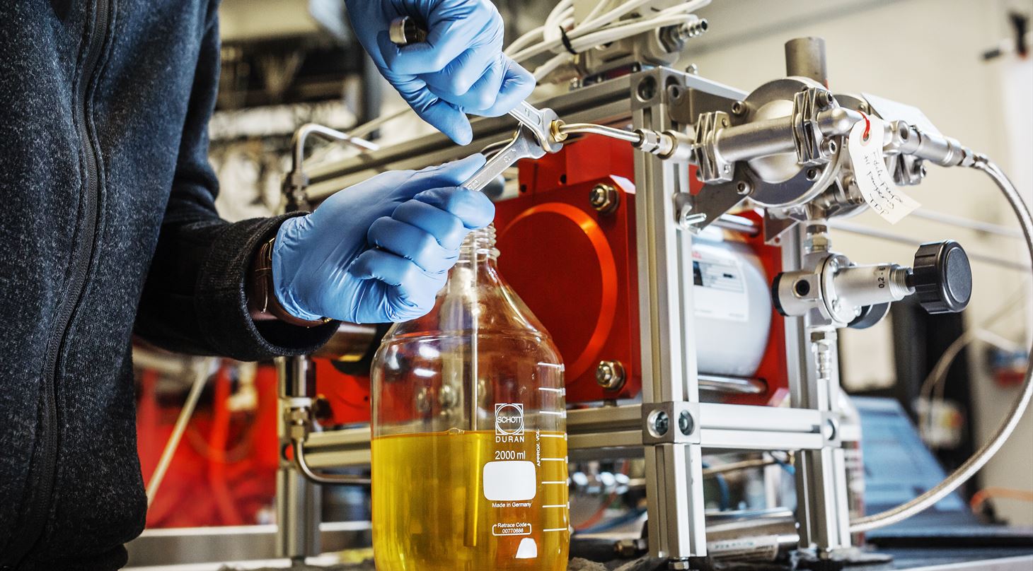 A man with blue nitrile gloves is using two combination spanners on a red machine. In the foreground, a flask with some yellow liquid is to be seen.