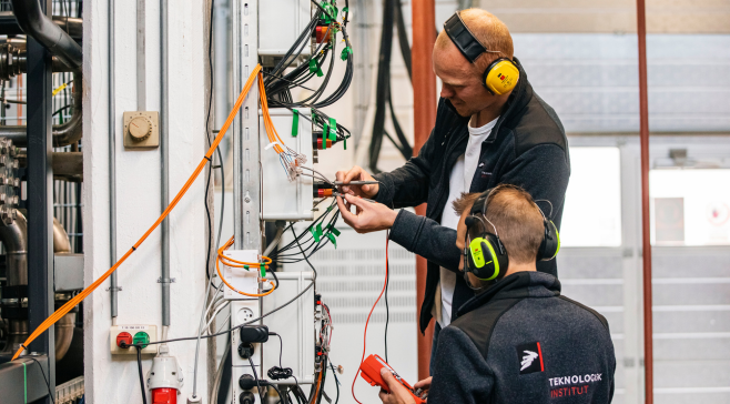 En tekniker med høreværn der arbejder ved et åbent el-panel og røranlæg i et testlaboratorium.