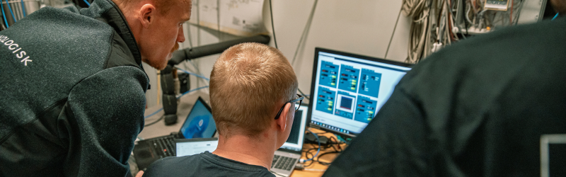 Tre teknikere i arbejdstøj der sidder samlet foran en bærbar computer i et testlaboratorium.