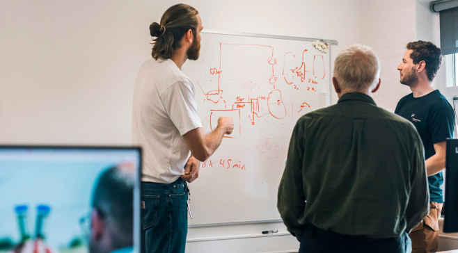 Tre mænd står samlet foran et whiteboard, hvor én af dem peger på og forklarer en håndtegnet teknisk tegning