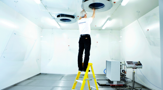 En tekniker står på en gul trappestige i et lyst testlokale og arbejder på en ventilationsåbning i loftet ved siden af en teknisk installation på gulvet.